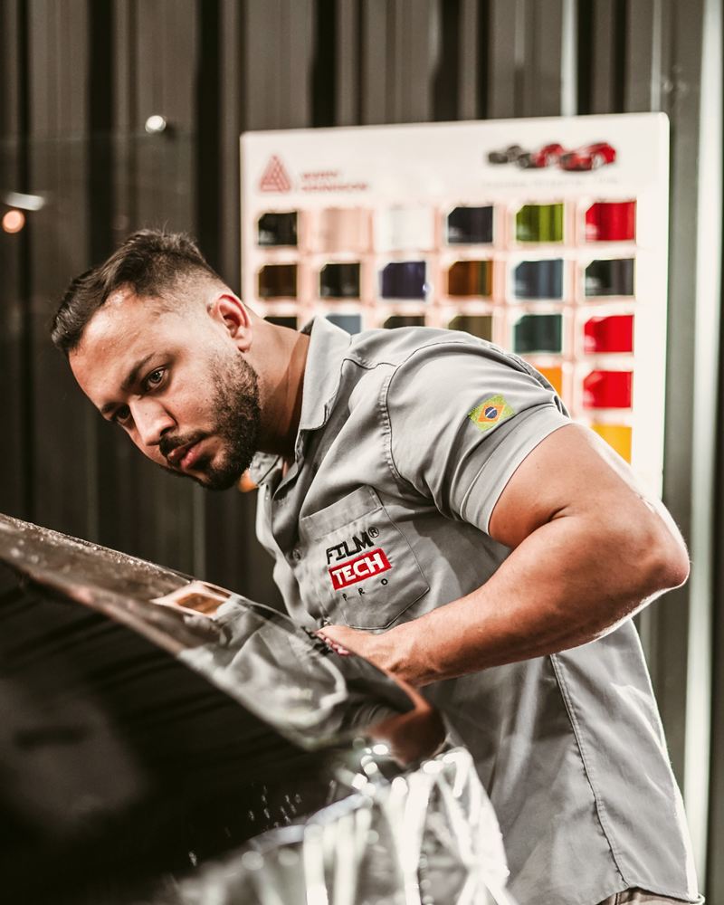 Pintor de uniforme cinza trabalhando na lataria de um carro preto, com o mostruário de cores ao fundo